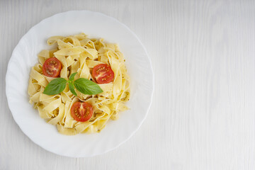 Top view of a plate full wth traditional italian fettucine pasta decorated with sliced tomatoes and basil leaves on white wooden background at kitchen. Image with copy space, horizontal orientation