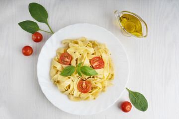 Top view of organic italian fettucine pasta decorated with sliced tomatoes and basil leaves in a plate surrounded by spinach and olive oil gravy boat on white wooden background at kitchen