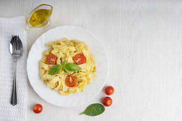 Top view of plate full of italian fettucine pasta decorated with basil leaves served with spoon and fork on napkin and olive oil gravy boat on white wooden background. Image with copy space