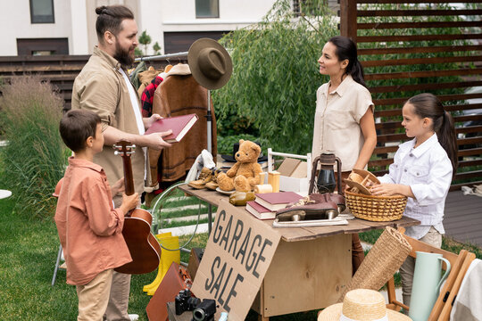 Young Bearded Man Buying Book And Boy Buying Guitar At Yard Sale Organized By Woman And Her Daughter