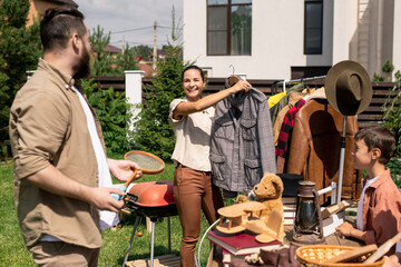 Positive young woman holding jacket on hanger and offering to buy it to husband at summer garage...