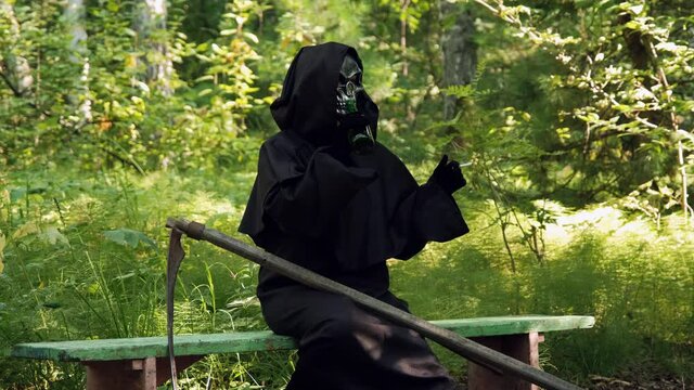 Grim Reaper with Cigarette in Hand Drinking Beer while Sitting on a Bench in a Park. Slow Motion. Life and Death, Another World and Halloween Concept