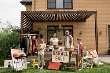 Portrait of positive young family with two kids waiting for customers at garage sale