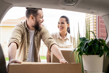 Excited young couple standing at car and taking moving box out of trunk while moving into new house