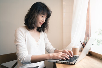 Young Asian woman working with laptop computer and drinking a cup of coffee. A happy woman with smiley face working from home. Social distancing concept