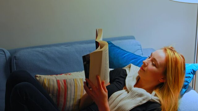 Smiling woman lying on safa and reading book. She is dreaming about novel.