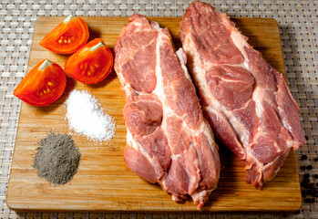 Two fresh pieces of pork meat chops on a wooden plank next to pepper salt tomatoes closeup concept of cooking meat dishes in restaurant food and outdoors
