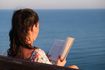 young woman reading a book outdoors with sea backgound and sunlight iluminating her face, ...