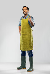 gardening, farming and people concept - happy smiling indian male gardener or farmer in apron and rubber boots showing thumbs up over grey background