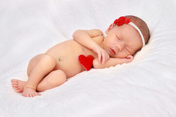 A newborn girl with a bandage on her head with red roses and a red heart is sleeping with her hand under her cheek.