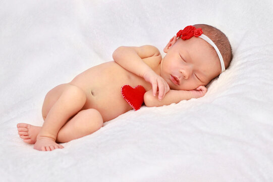 A newborn girl with a bandage on her head with red roses and a red heart is sleeping with her hand under her cheek.