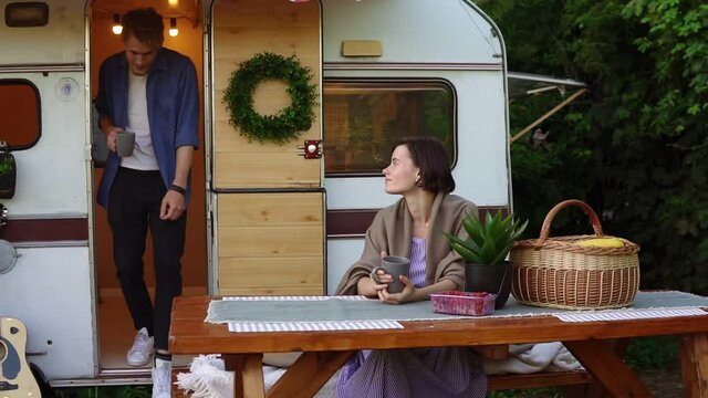 Portrait of cheerful couple outdoors near their trailer house. Girl sitting at wooden picnic table, drinking coffee and enjoying morning. Bearded man come out from huse with a mug