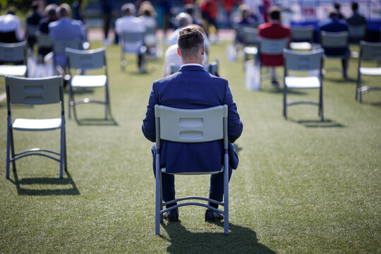 People Sit On Chairs Apart One From Another To Maintain The Social Distance During The Covid-19 Outbreak At An Outdoor Event On The Turf Of A Stadium.