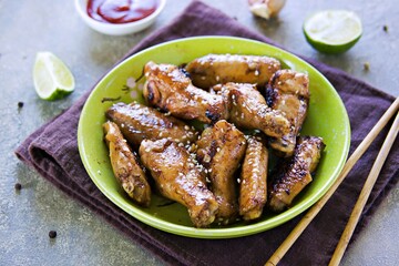 Fried Chinese style chicken wings sprinkled with white sesame seeds in a green ceramic bowl on a gray concrete background. Grill, barbecue recipes.