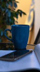 Evening coffee. Blue cup with coffee, open book and mobile phone, evening light, sunset, dusk. Atmospheric, cozy evening in the garden, green leaves, conversation. Warm light, blur bokeh.