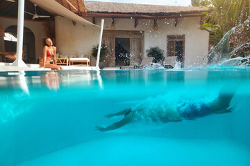 Couple At Pool. Man Underwater And Woman On Poolside Having Fun On Summer Vacation Or Honeymoon At  Luxury Resort. Diving Male Splashing Water And Female Looks At Him. Summertime At Tropical Villa.