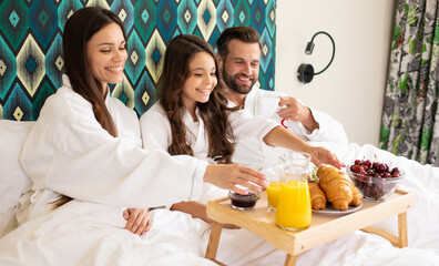 Obraz na płótnie Canvas Happy young beautiful family in bathrobes are taking morning breakfast in a luxury hotel room. Service in the room. Vacation and trip