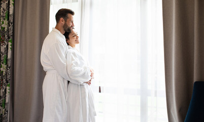 Tender and a beautiful young couple in love in white bathrobes in the luxury hotel or bedroom are standing near a big window and hugging each other