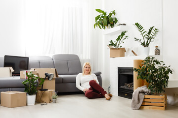 Happy young woman sitting on floor near moving boxes. Young woman moving to new home. Woman looking at camera and smiling