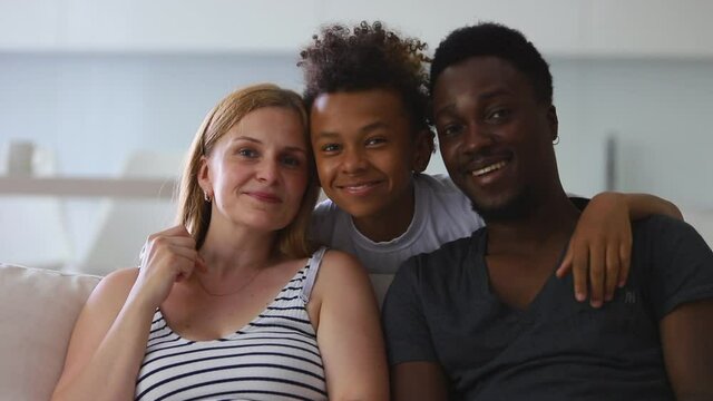 portrait of cute happy interracial family of mother, father and son. Spbi couple and mixed race boy sit on sofa and look at camera. teenager child hug embrace multiethnic parents.