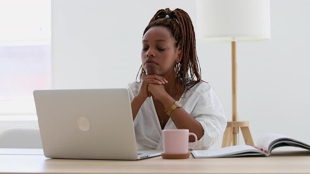 Young African American woman is working with laptop at table in light office spbi. Beautiful female looking at computer screen while sitting at desk in modern interior. Millennial person has work time