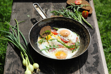 Rustic breakfast. Fried eggs with vegetables: tomatoes and asparagus beans in a pan with bread, herbs, onions on old wooden black background in nature. Summer, sun light. Background image, copy space