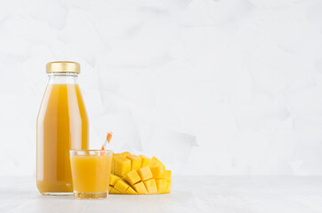 Summer orange pulpy mango in glass bottle mock up with straw, wine glass, fruit pieces on white wood table in light interior, template for packaging, advertising, design, branding product.