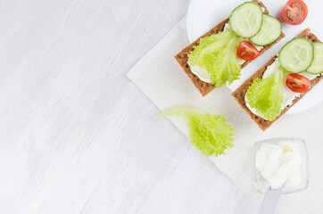 Healthy dietary breakfast of crisps rye flat toast with fresh vegetables - green salad, cucumber, tomato, cream cheese on white wood background, top view, copy space.