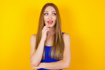 Dreamy female with thoughtful expression, looks to the camera, keeps hand near face, bitting a finger thinks about something pleasant, poses against gray wall.