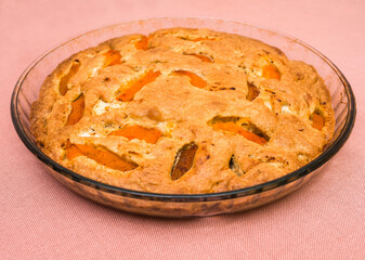 A large, round baked apple and apricot pie stands in a glass form on a table with a pink tablecloth. Delicious, fresh dessert.
