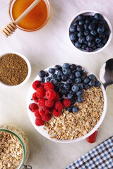 Top down view of fresh muesli breakfast on top of the kitchen table. White bowl with flakes, blueberry, raspberry and honey on white tablecloth.