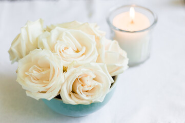 Beige roses in the light blue bowl with the candle on the background on the white fabric