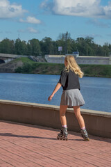 A young girl with blond hair, in a black T-shirt and a plaid skirt, is rollerblading along the city embankment on a summer day. The cityscape in the background is blurred.