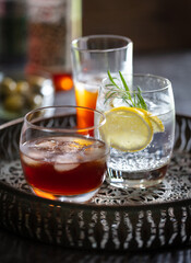 Glasses of brandy, gin & tonic and vermouth on drinks tray. Olives and bottle in background.