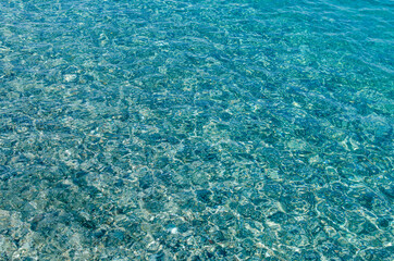 Natural background of transparent azure sea water and bottom with rocks. A close-up view of the pebbles on the beach. Blue bottom of the Mediterranean sea with small stones through clear water