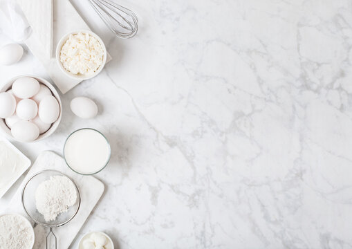 Fresh dairy products on white marble background. Glass of milk, bowl of flour, sour cream and cottage cheese and eggs. Steel whisk. Top view. Space for text