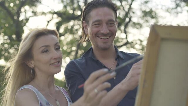 Close-up of laughing Caucasian couple of artists applying strokes on canvas and talking. Portrait of loving man and woman painting picture outdoors together. Positive painters creating artwork.