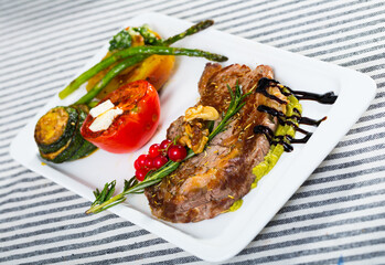Appetizing grilled veal loin with guacamole and baked vegetables served on ceramic plate on striped tablecloth..