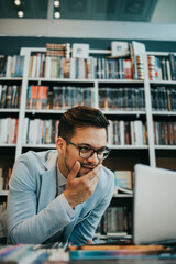 Young nice looking student or writer sitting in college library next to the window and working on his laptop. Natural light with strong and deep shadows. Wall with books in the background.