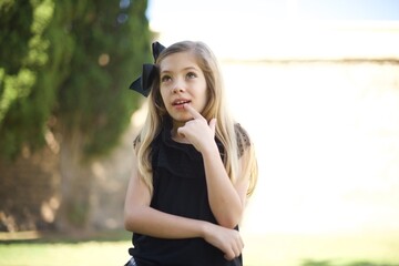 Dreamy female with thoughtful expression, looks to the camera, keeps hand near face, bitting a finger thinks about something pleasant, poses against gray wall.