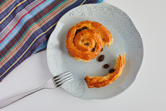 Raisin Bun Cut In 2 Pieces On A Plate With A Fork On The Side, Shot On White Table With Tablecloth Folded At The Corner In Top View