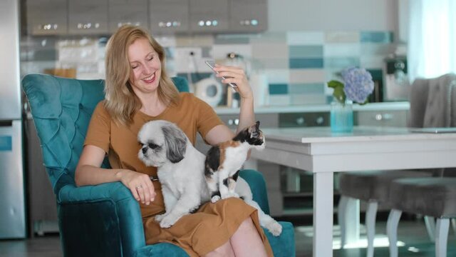 Young American woman taking selfie photo with pets sitting in armchair at apartment room rbbro. Attractive female touching cat, dog and making picture with smile while holding smartphone in hand at