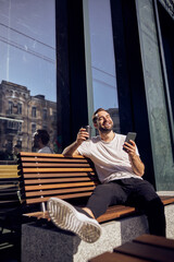 Fototapeta na wymiar Fun man with smartphone and coffee sitting on a bench closing his eyes outdoor