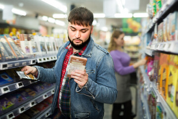 Attentive bearded guy choosing chocolate on shelves in grocery store.