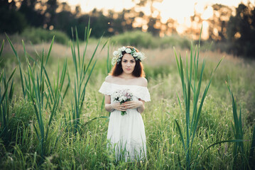 Caucasian happy girl in a white dress with a bouquet of flowers and a wreath in the summer