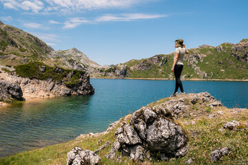 Naklejka na ściany i meble Young woman wearing a hat admiring the scenery of a lake surrounded by mountains in Somiedo Natural Park, Asturias.