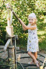 A little girl, a child in a colored sundress and a kerchief, stands and works, helping adults, agronomists near a manual metal pump with a well in a village outdoors.