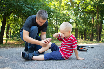 A four-year-old boy fell off a scooter and broke his knee. Dad provides first aid by disinfecting the wound and applying a plaster.