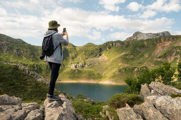 Naklejka na ściany i meble Young woman taking a picture of a lake with her mobile phone in Somiedo Natural Park, Asturias.