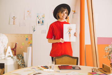 Pretty smiling girl artist in hat stands with drawing in art studio. Photography for ad of art...